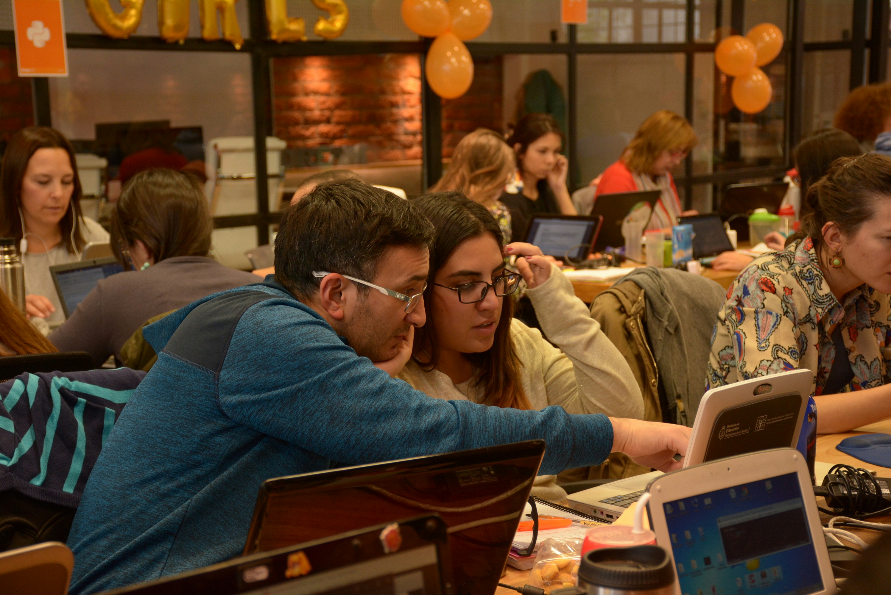 A mentor and mentee reviewing code together in a collaborative learning environment, surrounded by laptops and teams in a tech workshop, illustrating mentorship and leadership development in action.