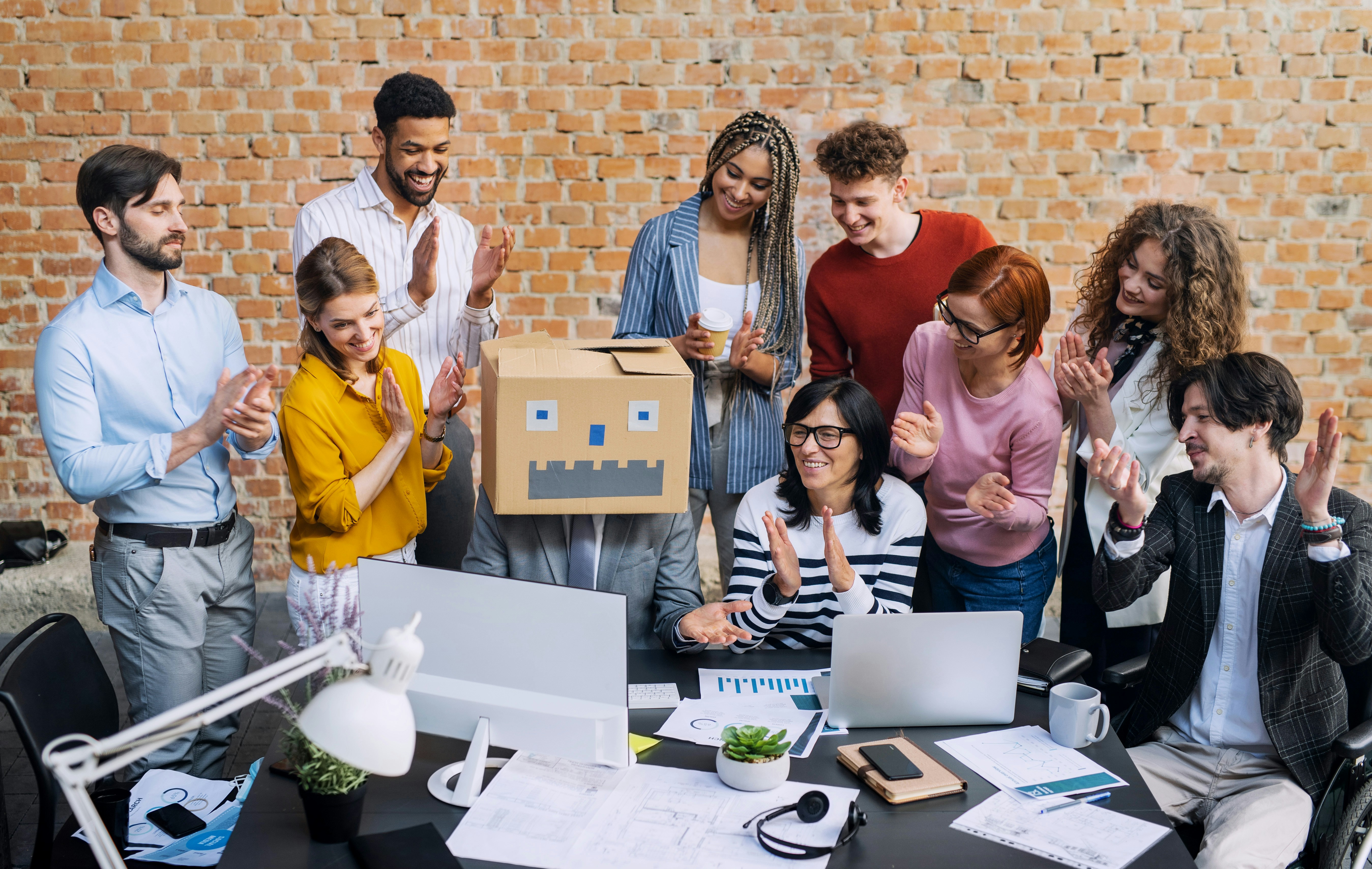 Team applauds a colleague in a robot head, symbolizing AI agents in the workplace.