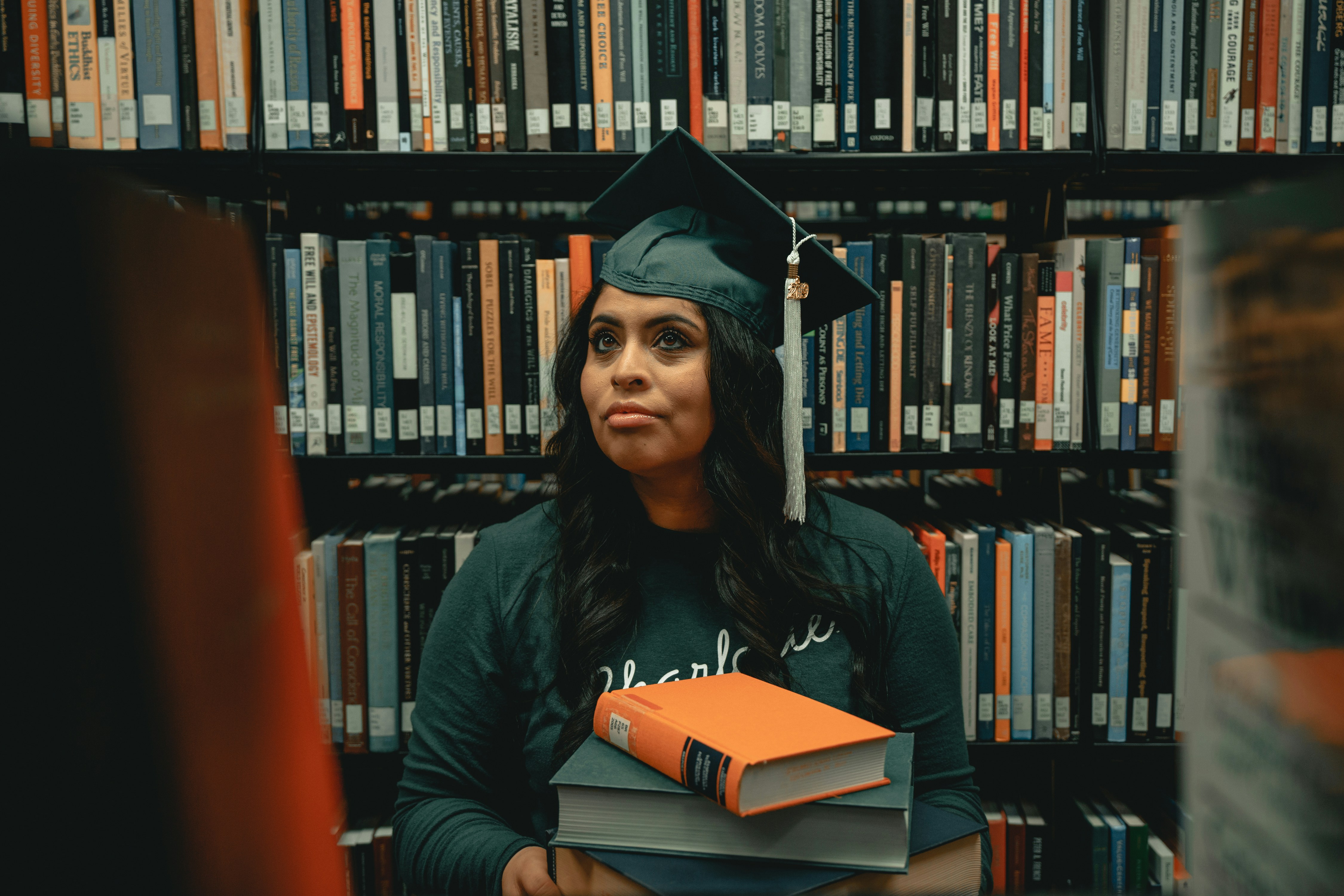 woman in a graduation cap and gown stands in a library, holding a stack of books, symbolizing the value of an AI Certificate Program in advancing education and career opportunities.